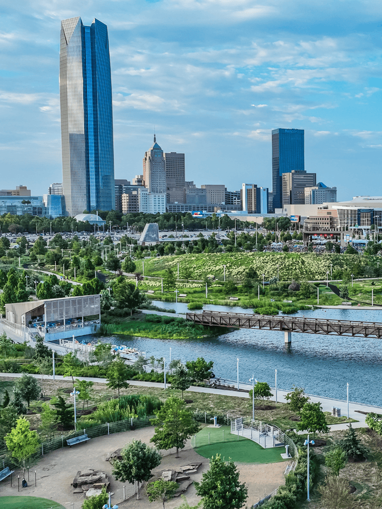 Modern city skyline with high-rise buildings and lush park with walking paths and river in the foreground.