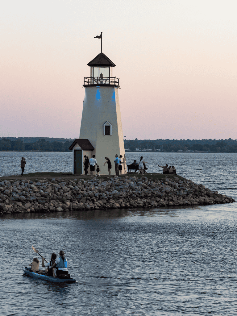 Lighthouse on a small rocky island with people enjoying water activities at sunset.