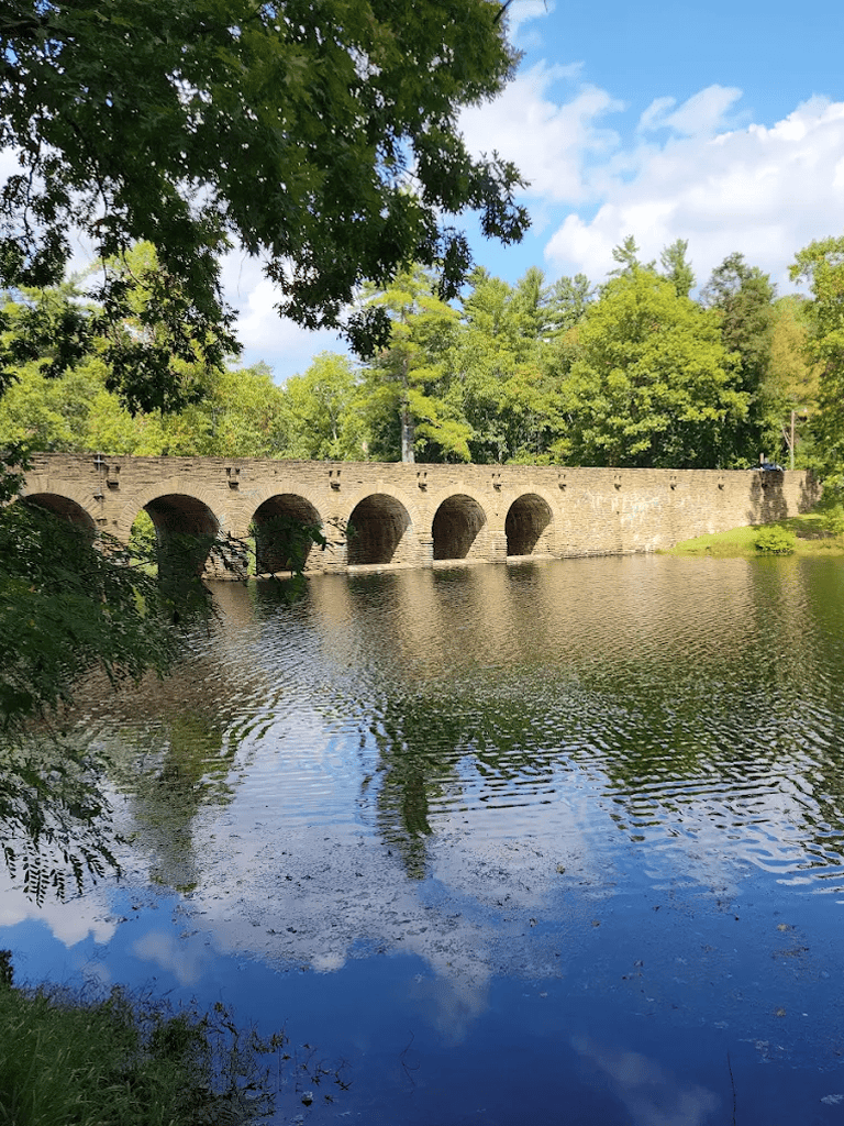 Stone bridge over river with lush green trees and blue sky in the background, scenic outdoor location.