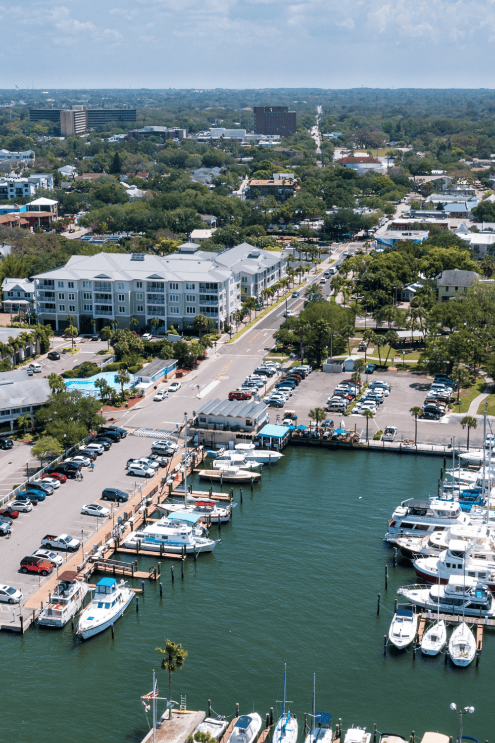 Boats docked at marina in sunny coastal city, colorful waterfront, lush greenery, residential and office buildings.