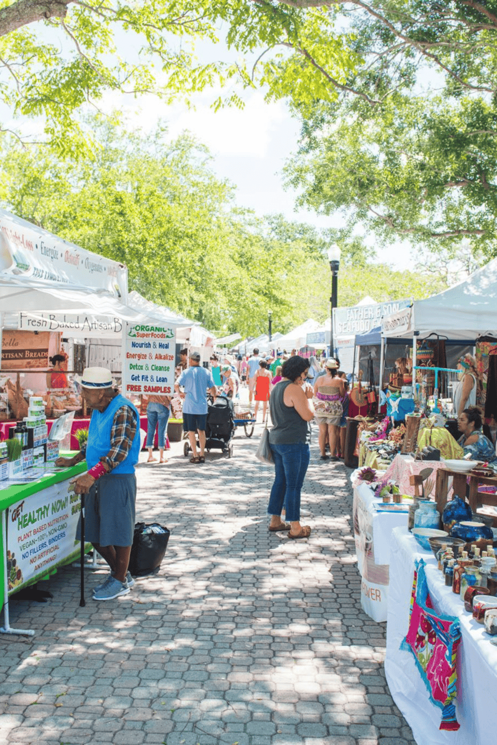 Vibrant outdoor farmers market with various artisan vendors and lush green trees in the background.