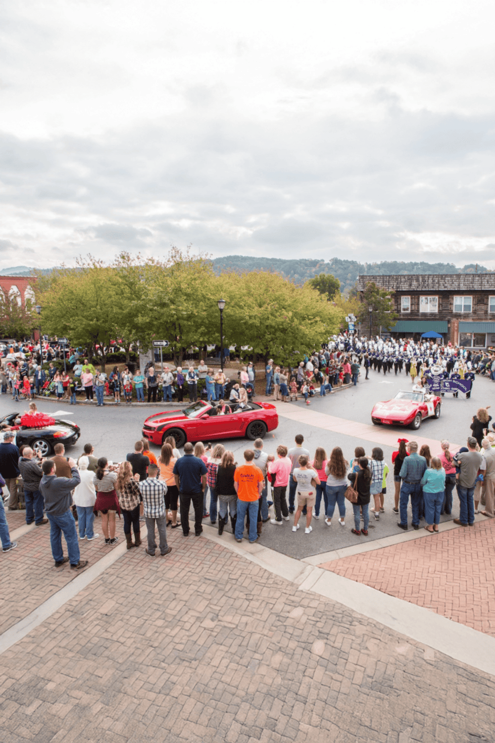 Vintage car parade in a small town street with crowd and parade band, scenic mountain backdrop.