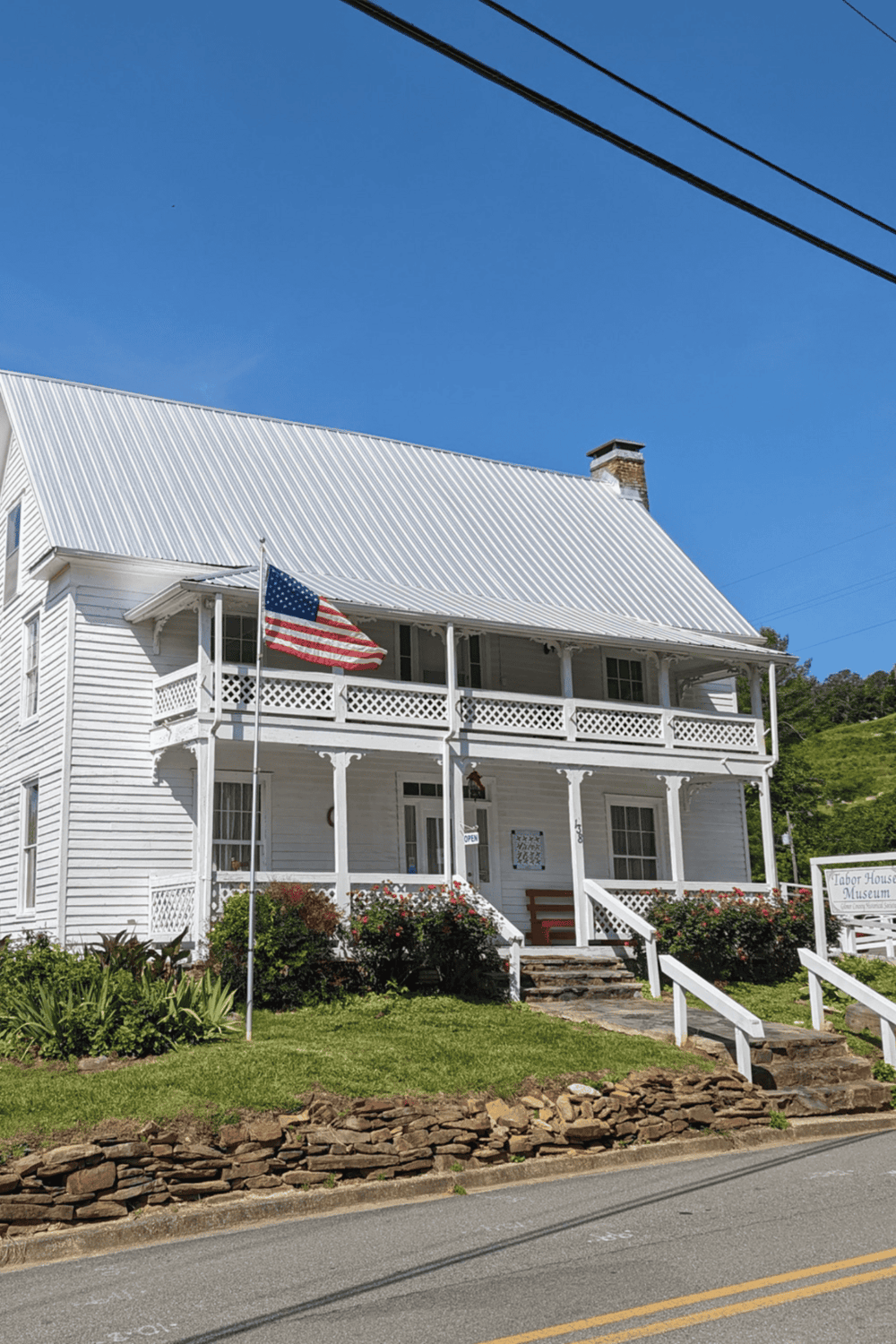 A white historic house with American flag, featuring a porch and lush greenery under clear blue sky.
