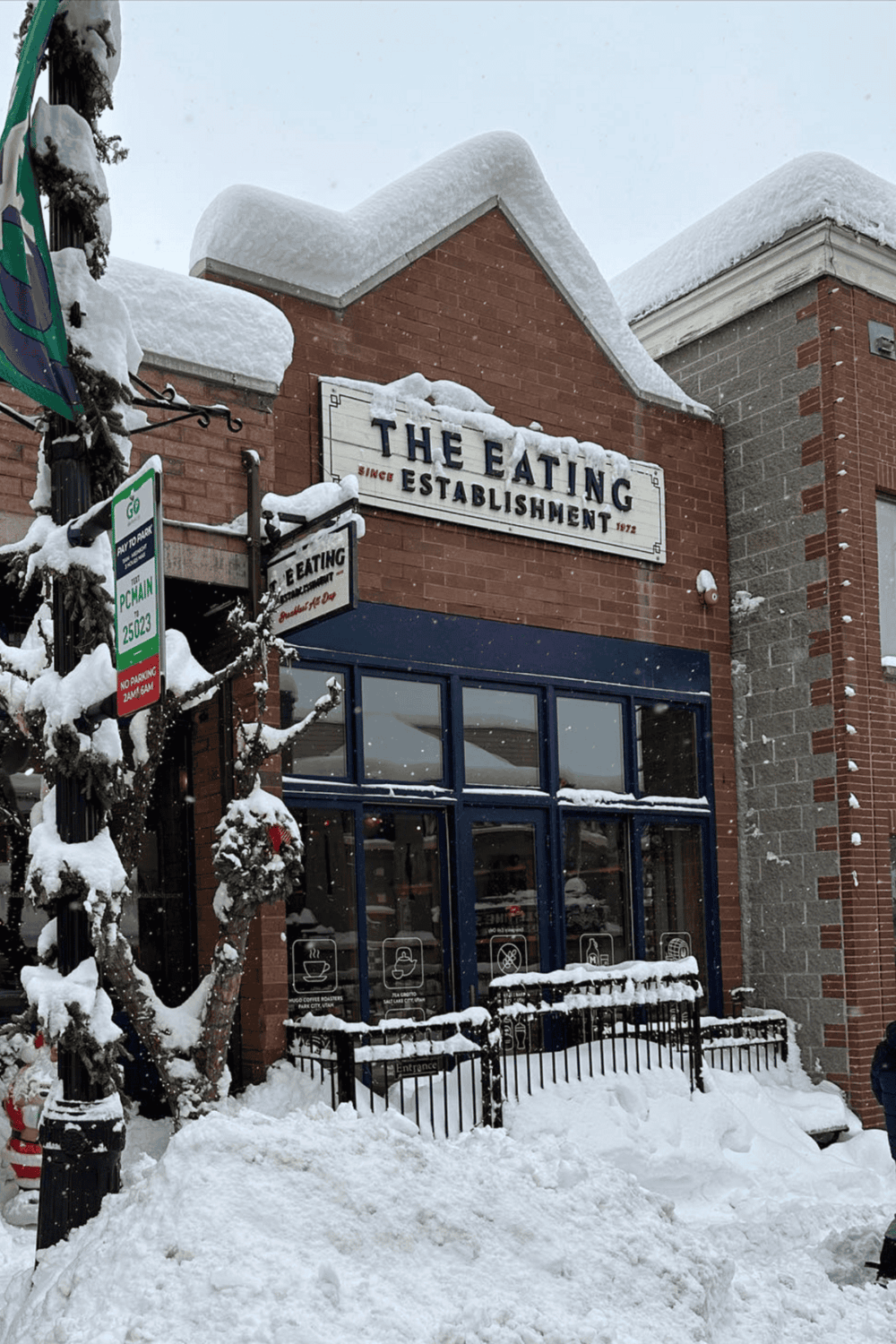 Snow-covered restaurant exterior with "The Eating" sign, winter scene, snow piled on roof and ground, inviting dining atmosphere, QuestForDirections SEO.