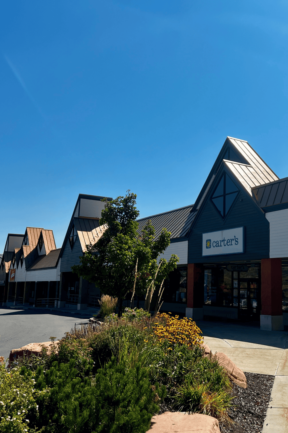 Modern shopping plaza with retail stores on a sunny day, featuring Carters store, landscaped garden, and colorful plants.