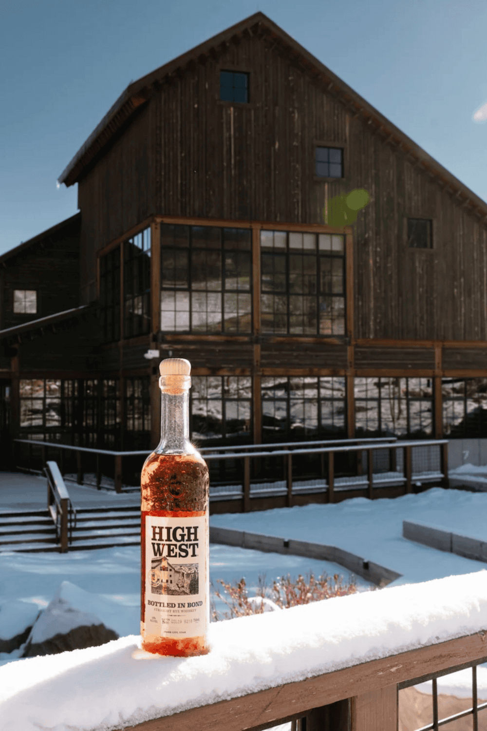 Bottle of High West whiskey on snow-covered balcony with rustic barn background.