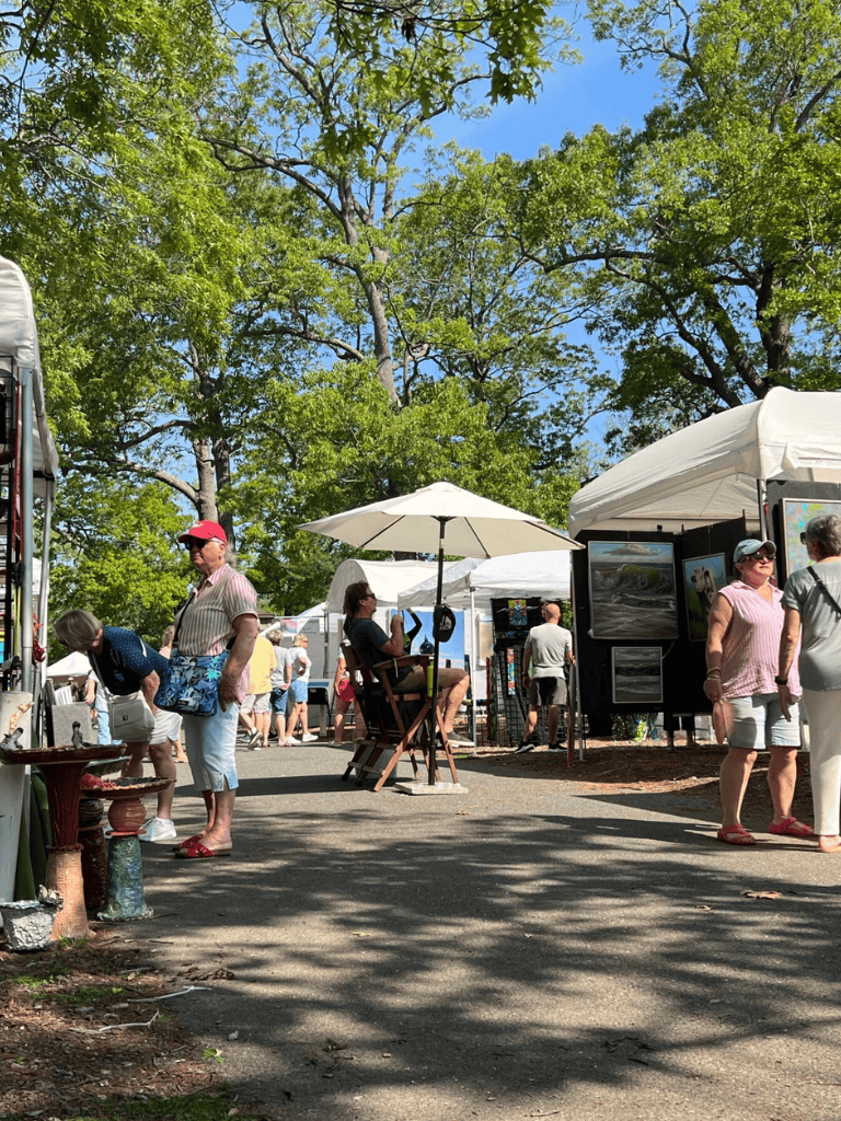 Colorful outdoor art fair with vendors, shoppers, trees, and bright blue sky.