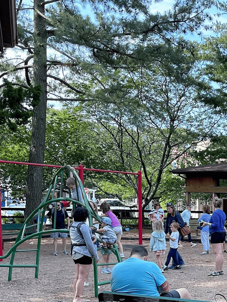 Child-friendly playground with children playing and adults supervising, lush green trees, outdoor community space, family fun, park recreation.