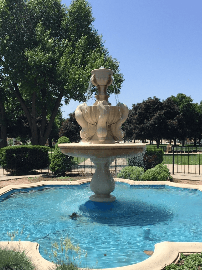 Elegant stone fountain in a lush park with green trees and a clear blue sky.