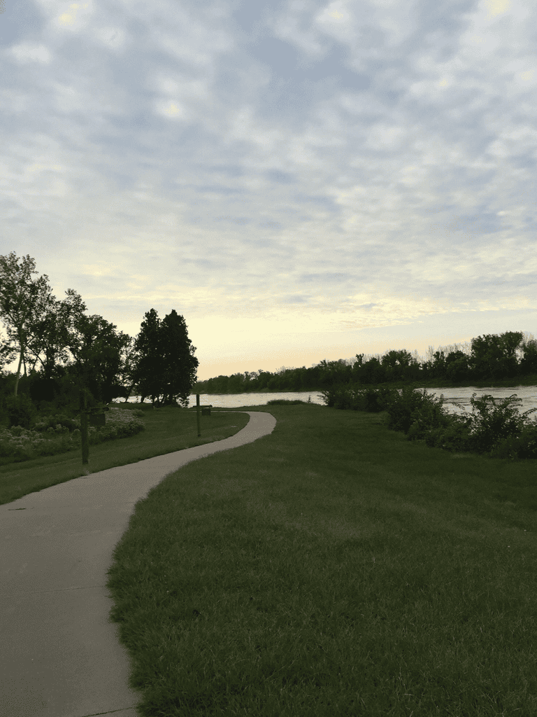 Serene riverside park with walking trail and lush green trees, perfect for outdoor exploration and relaxation.