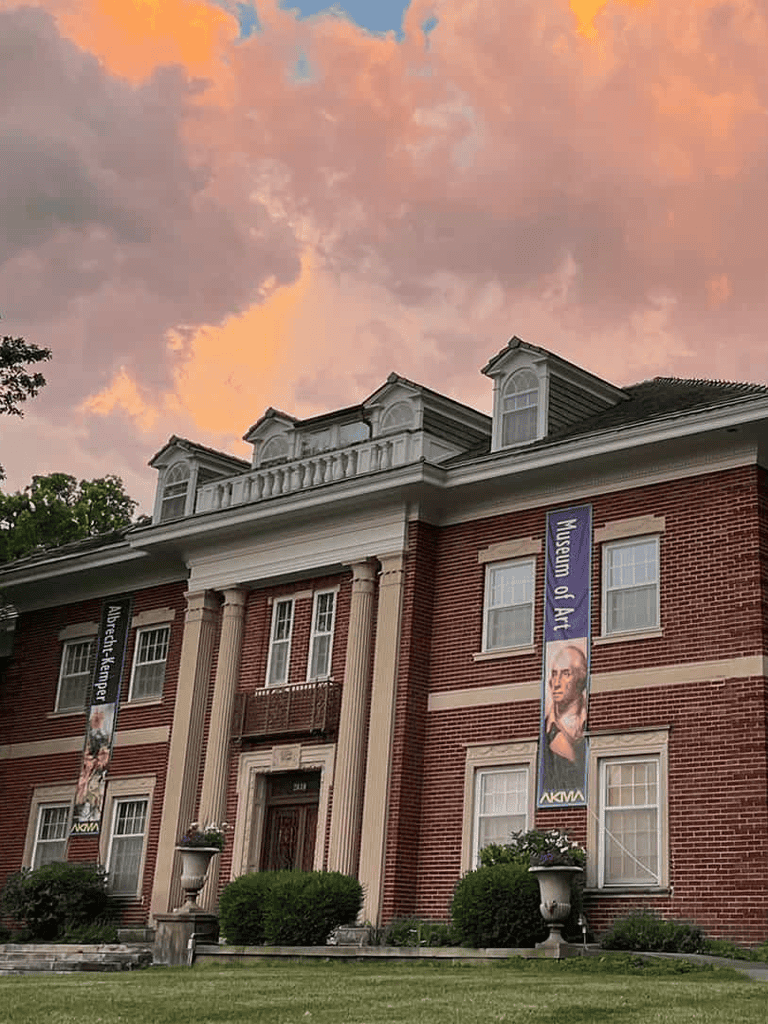 Elegant historic brick museum building with flags, lush greenery, and colorful sunset sky.