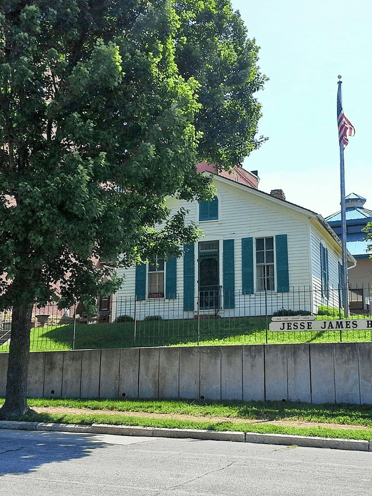 Old white house with blue shutters and American flag, located on Jesse James Highway, in a green neighborhood setting.