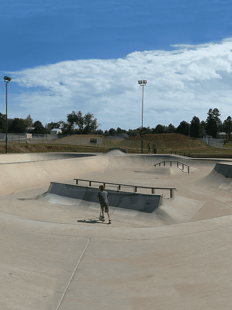 Skateboarder at concrete skate park with ramps and rails under blue sky.