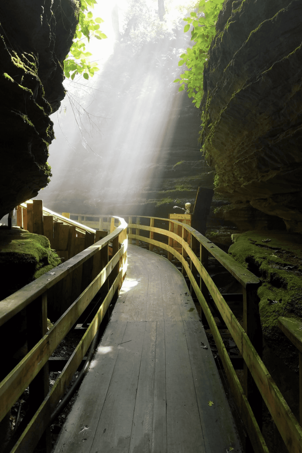 A peaceful forest waterfall with a wooden walkway and sunlight streaming through trees.