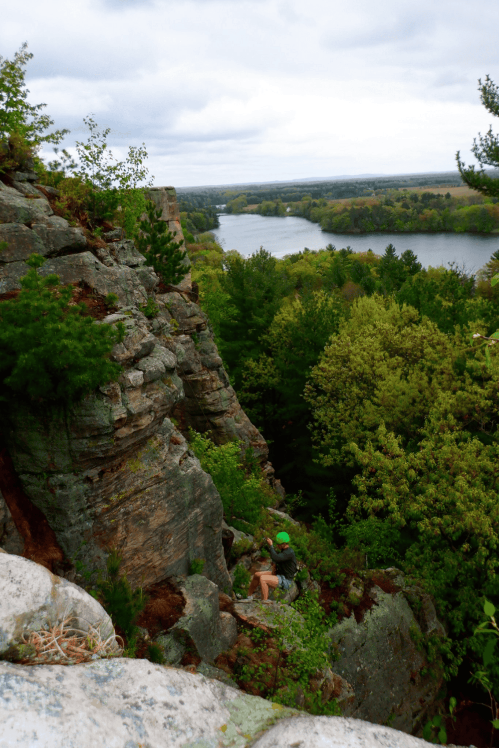 Abandoned rock formation overlooking lush river valley with hiker in green helmet exploring forest trail.