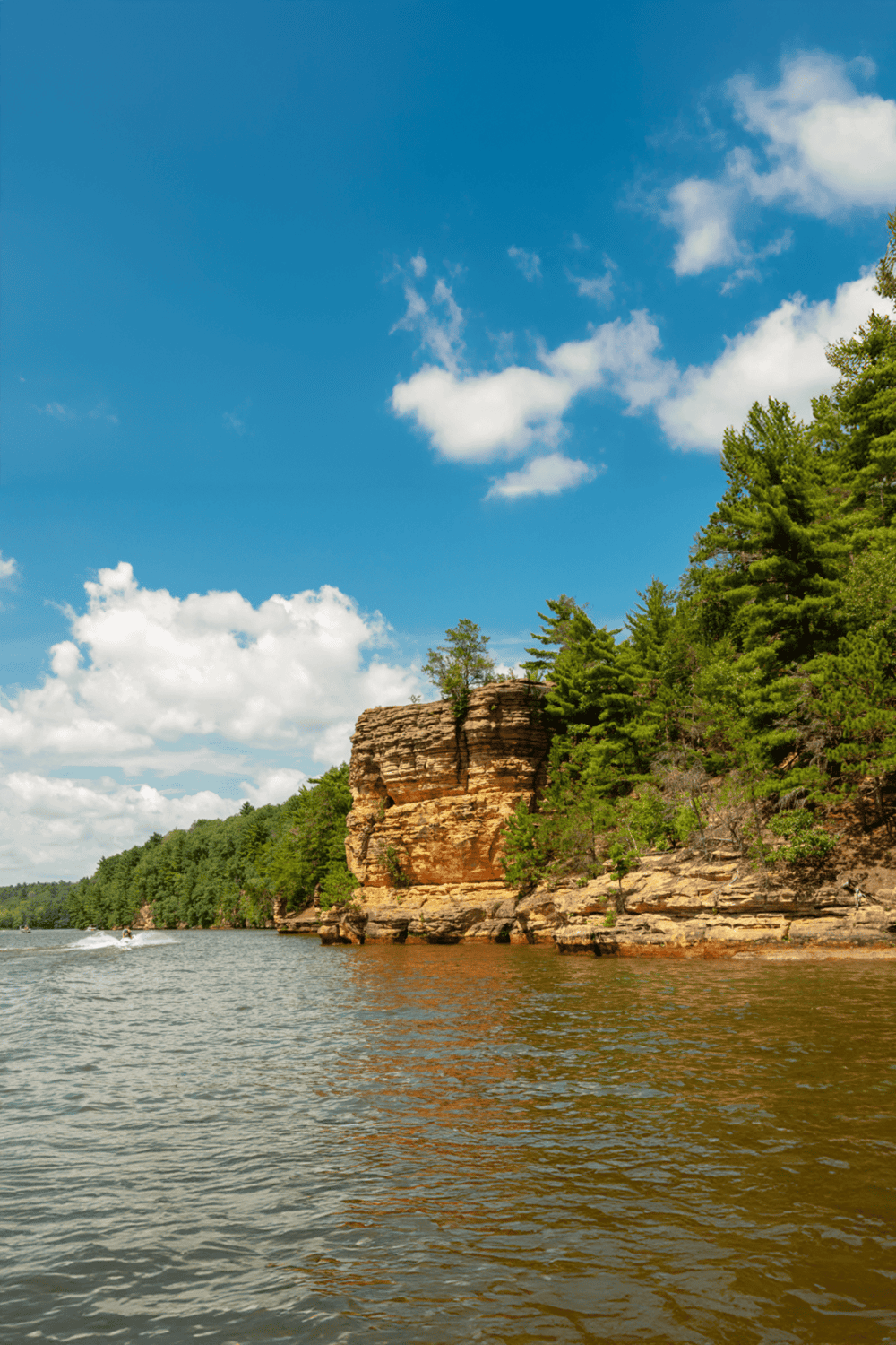 Vast river with rocky cliffs and lush green trees under a bright blue sky with scattered clouds.