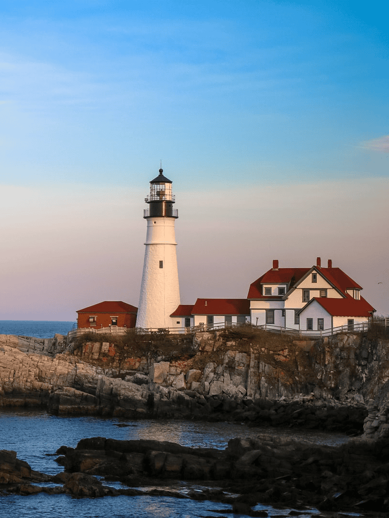 Sunset lighthouse on rocky coast with white house, coastal scenery, and calm ocean waters.