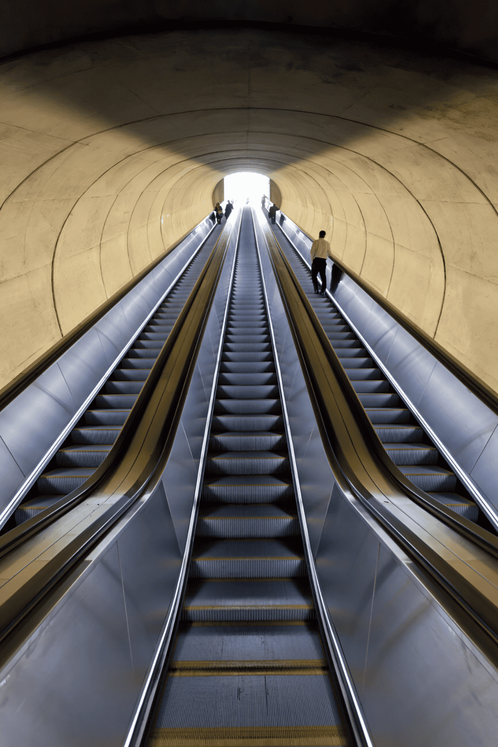 Escalator in a modern underground tunnel with curved walls, bright light at the exit, and people traveling.