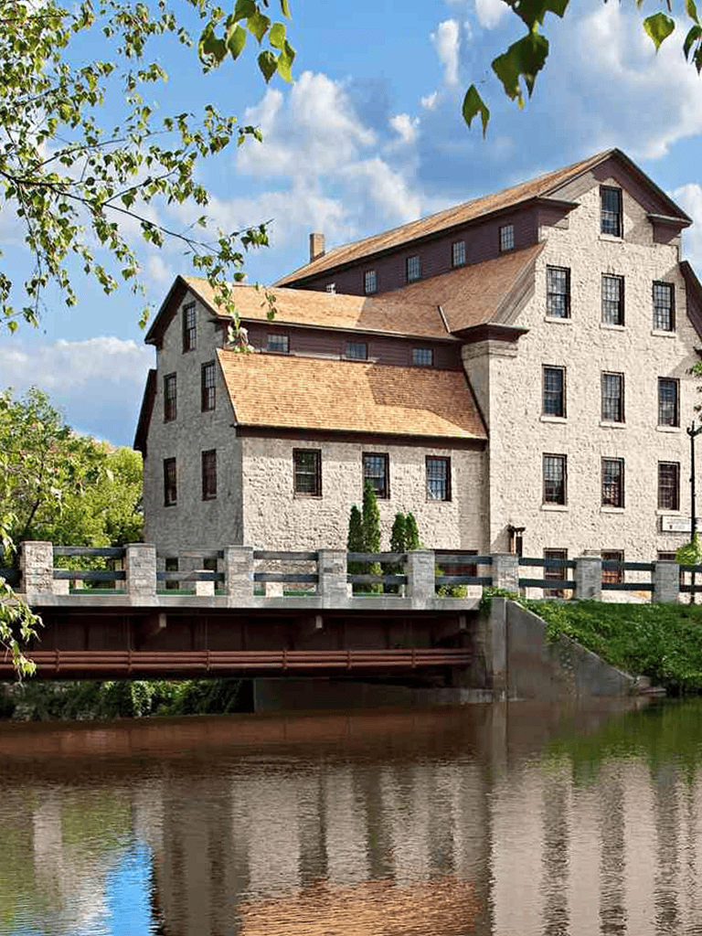 Historic stone mill building on riverbank with lush green surroundings and blue sky.