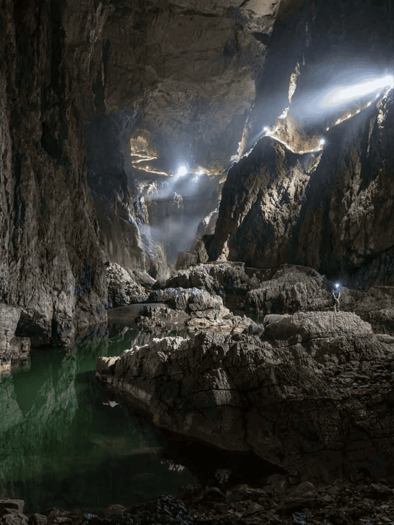 Ancient cave with sunlight beams shining through, reflecting on green water inside a rugged stone cavern.