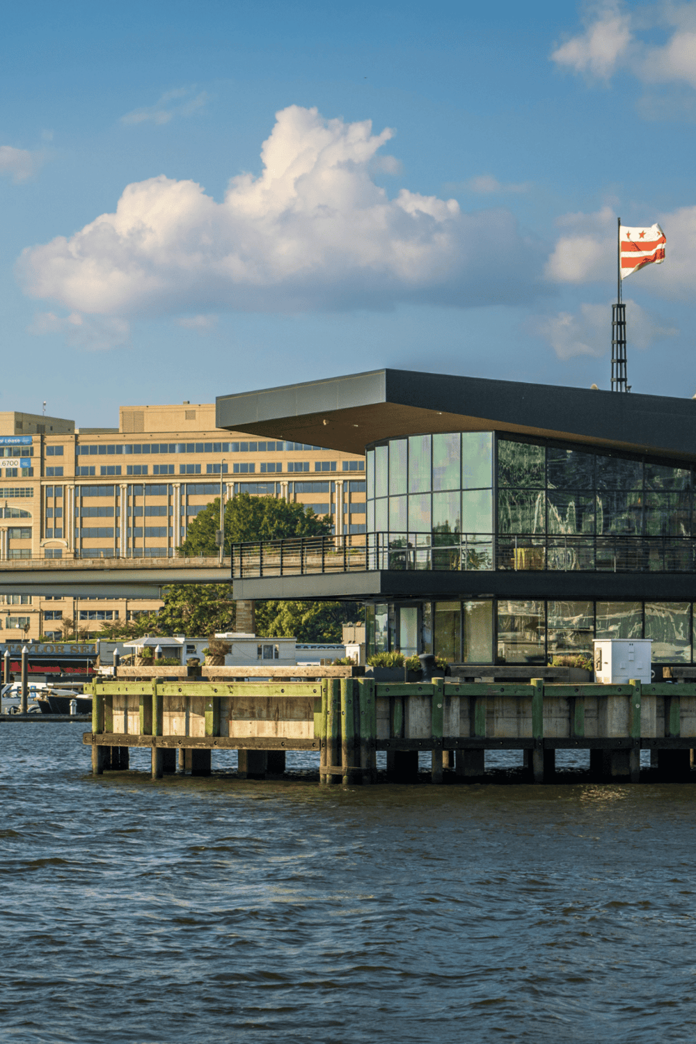 Modern waterfront building on water with cityscape background and flag flying in the sky.