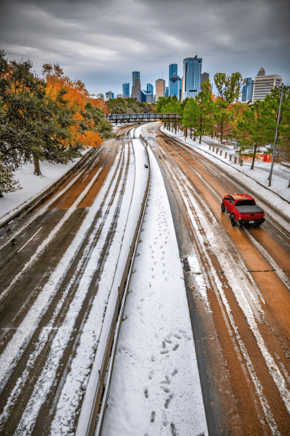 Snow-covered city highway with a red truck, overlooking downtown skyscrapers, during winter.