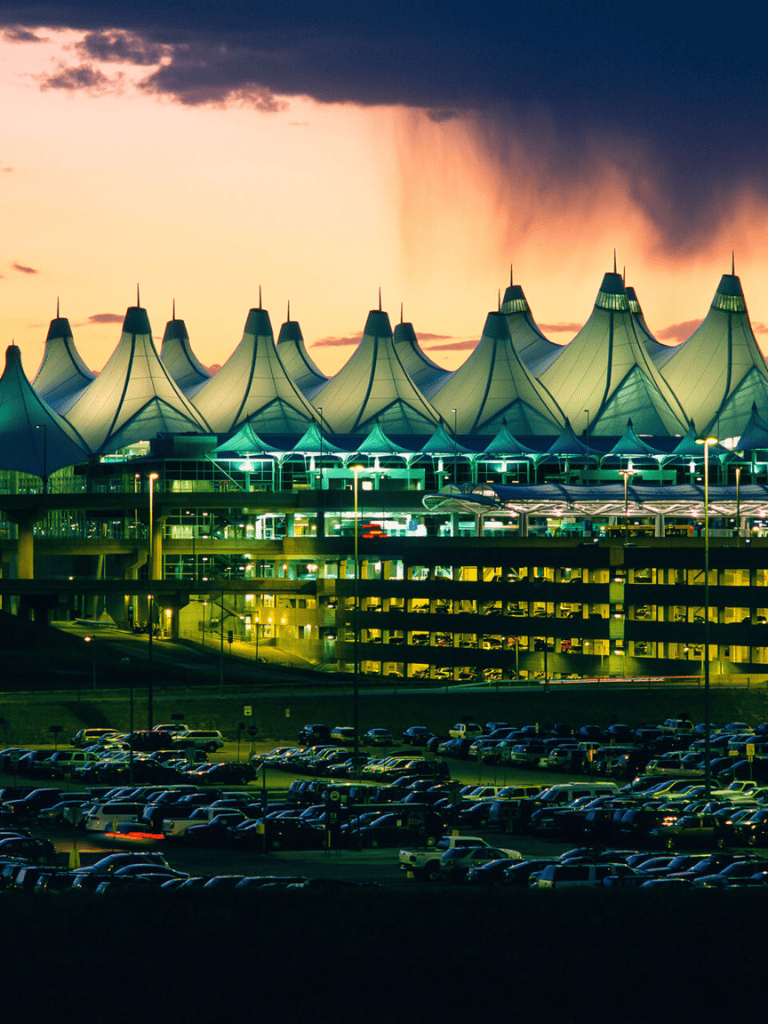 Modern airport terminal with distinctive tent-like roof structure at dusk, illuminated parking lot in foreground.