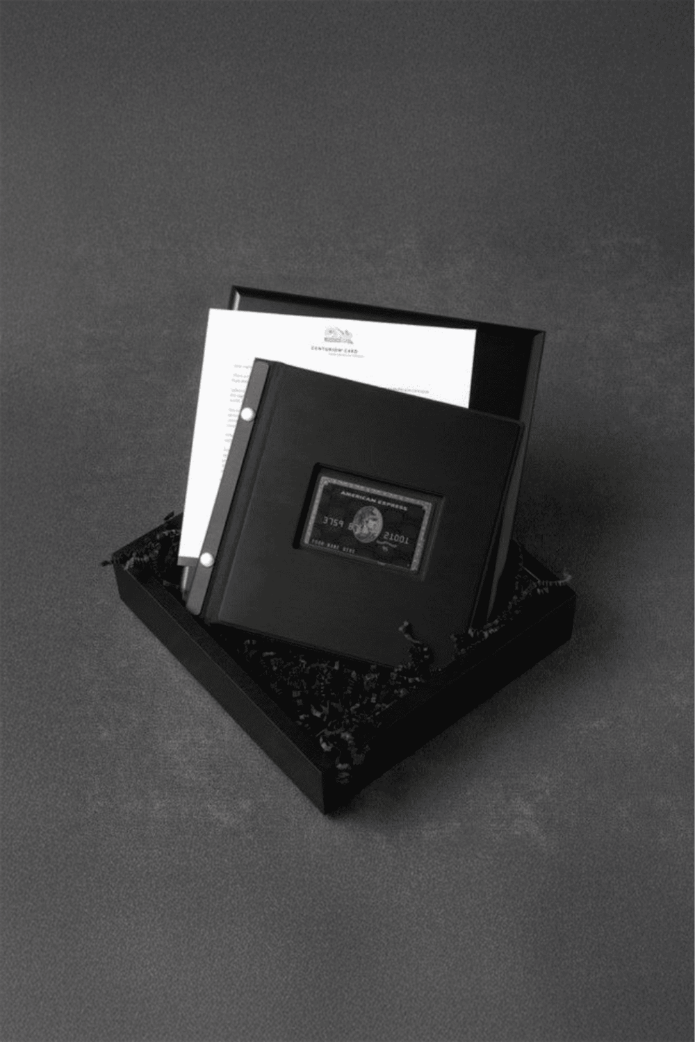 A black and white image of a branded gift box containing a leather checkbook holder, documents, and a credit card.