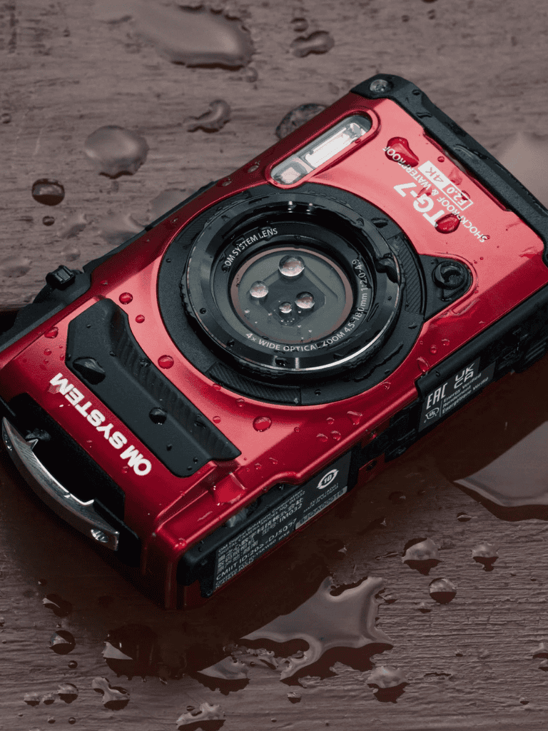 Waterproof red digital camera on wet wooden surface with water drops, ideal for outdoor photography.