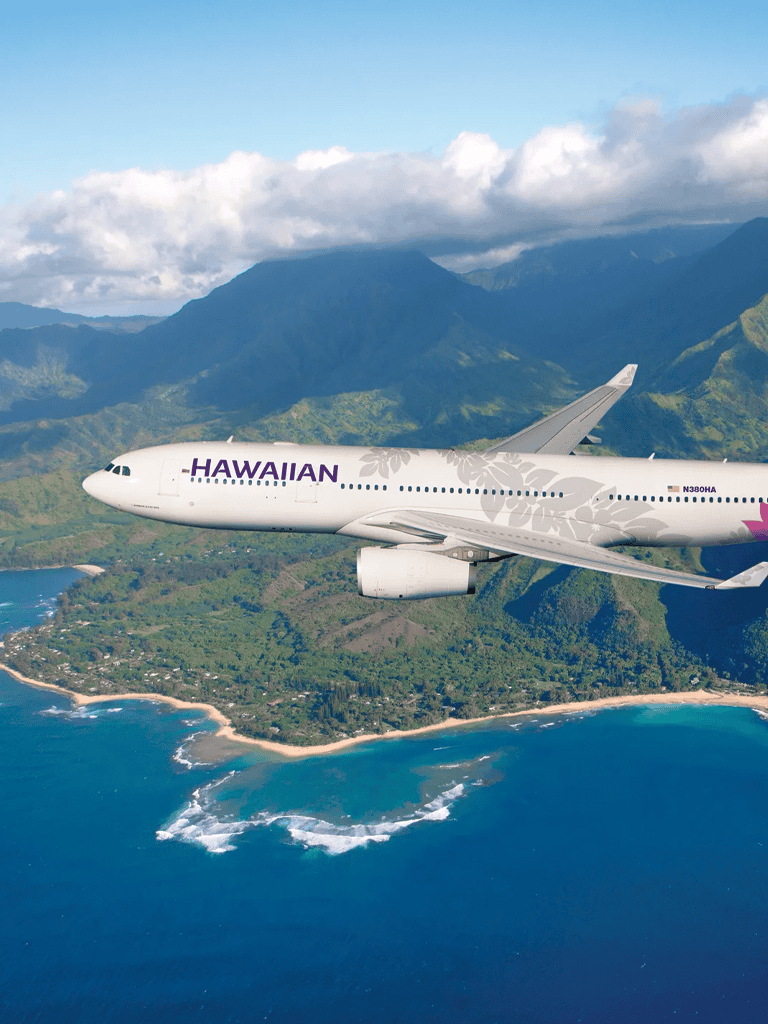 Hawaiian Airlines airplane flying over lush Hawaiian coastline and mountains.