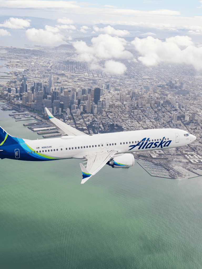 Alaska Airlines airplane flying over city skyline and water.