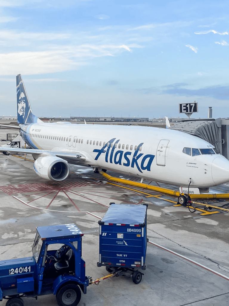 Alaska Airlines airplane at airport gate with jet bridge and ground service vehicles.