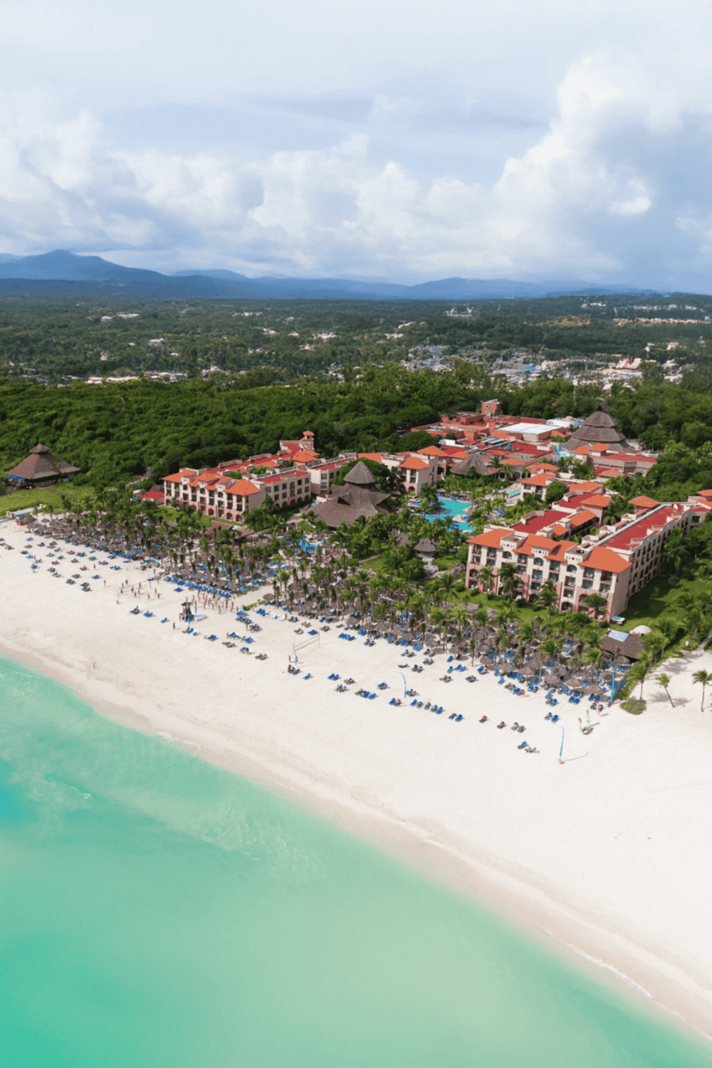 Beach resort with white sand and turquoise water, lush greenery, and scenic mountain views in the background.