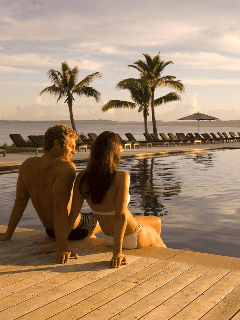 Relaxing tropical beach scene with couple by pool at sunset, palm trees, lounge chairs, and ocean view, perfect for travel and vacation SEO.