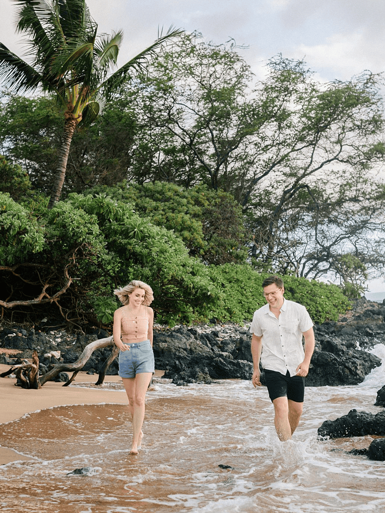 Relaxed couple walking on rocky beach with lush greenery in the background, tropical vacation scenery.