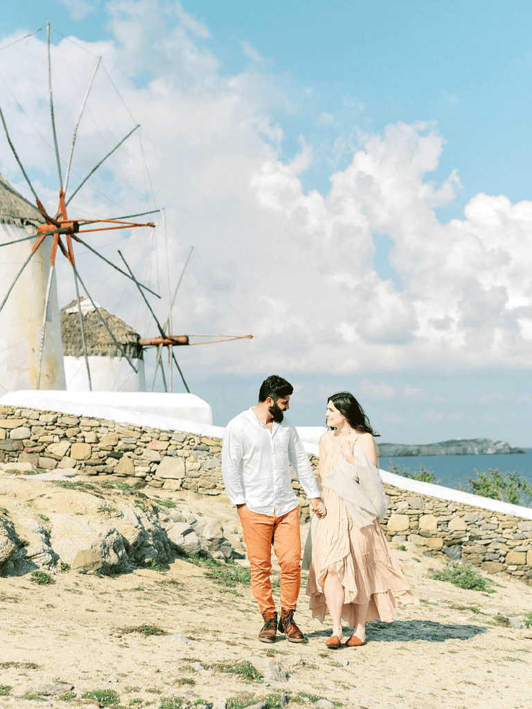 Romantic couple walking hand in hand on coastal path with windmill and blue sky.