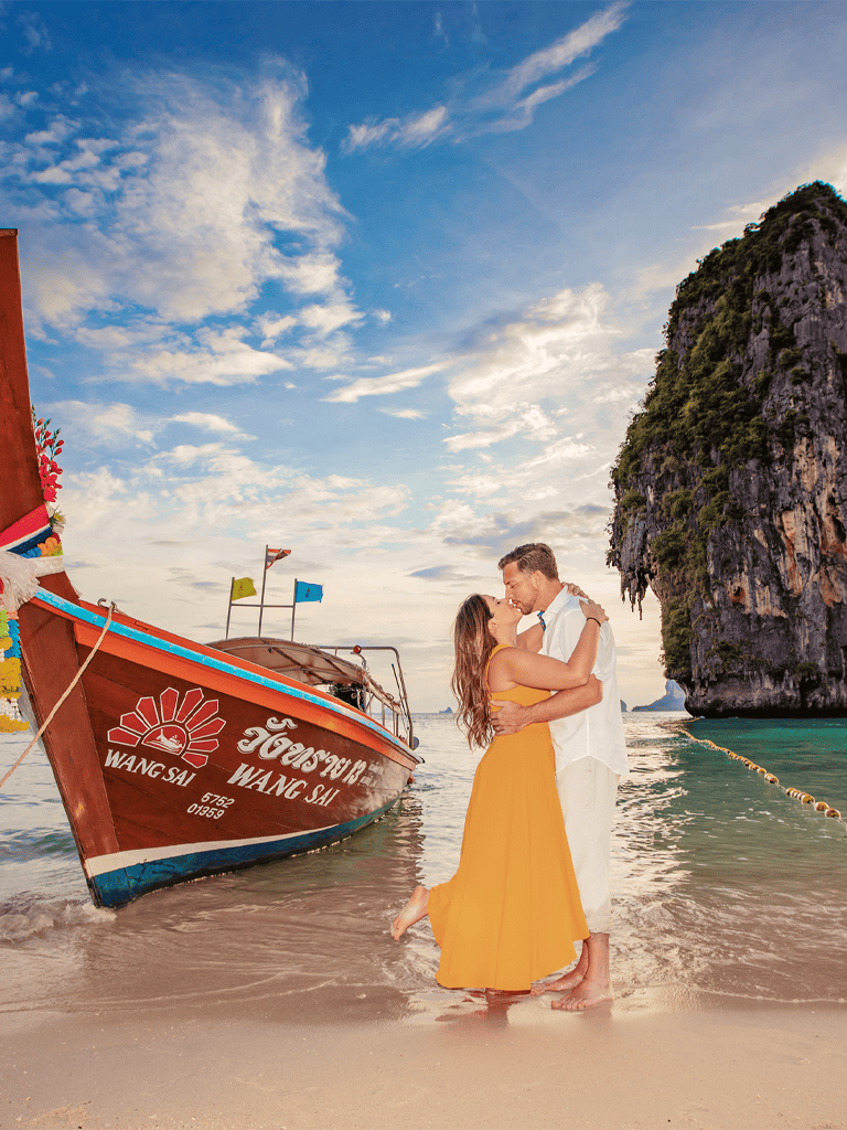 Romantic couple embracing on tropical beach with boat, limestone cliffs, and sunset sky.
