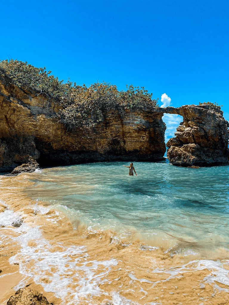 Bright blue sky over natural rock arch on tropical beach with turquoise water and sandy shoreline.