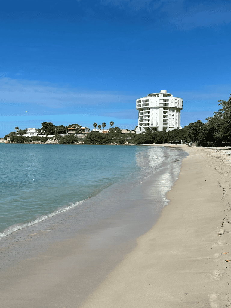 Relaxing beach with modern waterfront hotel in the background, sunny day, clear waters, and soft sandy shoreline.