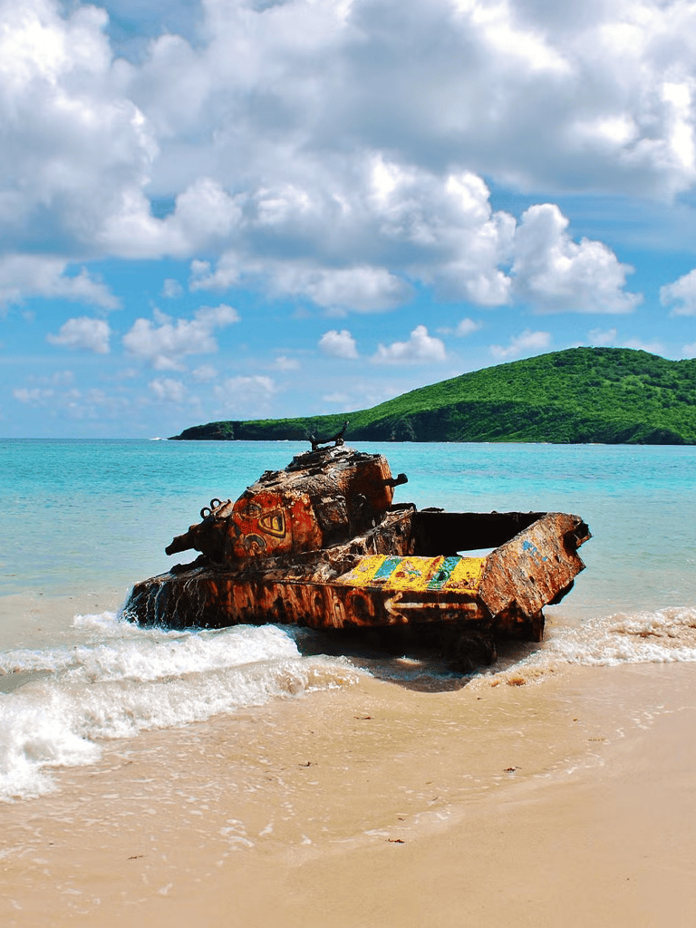 Wrecked military tank on tropical beach with green island and cloudy sky backdrop.