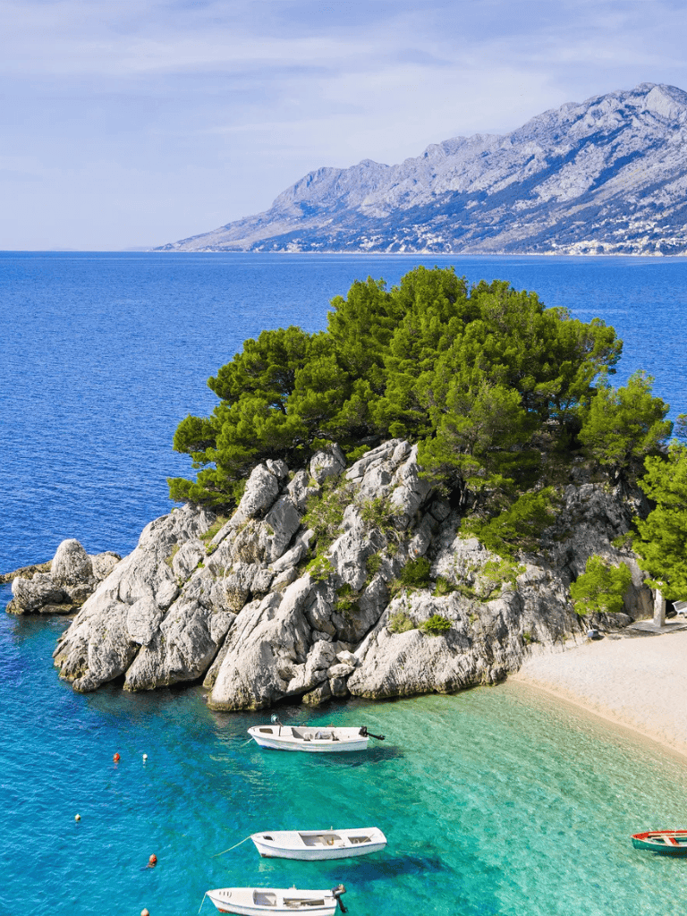 Stunning coastal landscape with rocky island, green trees, and boats in crystal-clear waters.