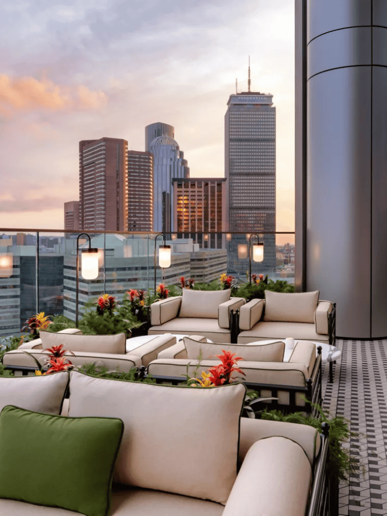 Outdoor rooftop lounge with city skyline view at sunset, modern furniture, and floral decor.