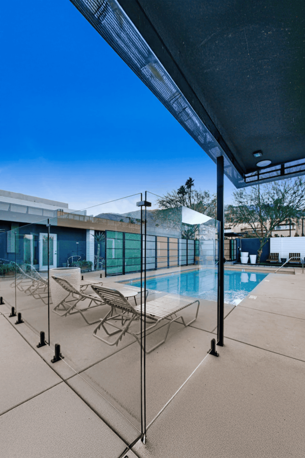Modern hotel pool area with glass fencing, lounge chairs, and sunny sky, perfect for relaxation.