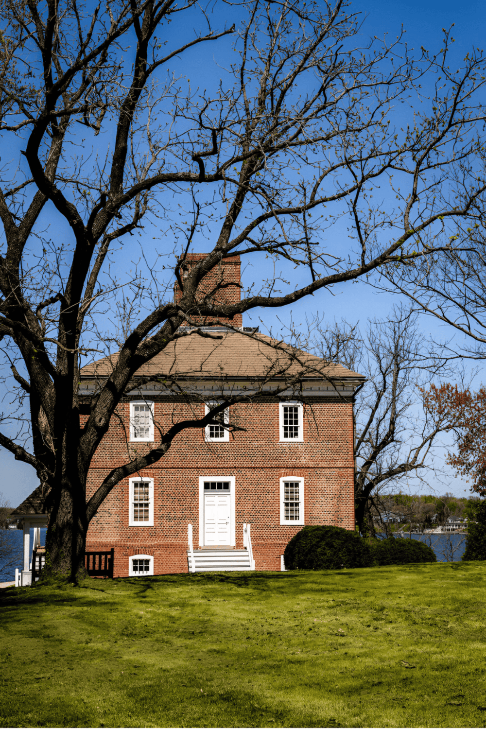 Old brick house overlooking a calm river with leafless trees and green grass in foreground.