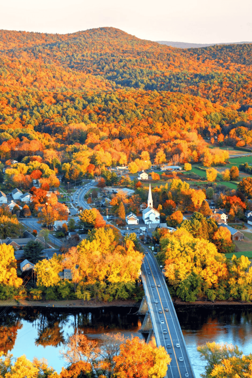 Colorful autumn landscape with a small town, church, bridge, and vibrant fall foliage in a scenic rural area.