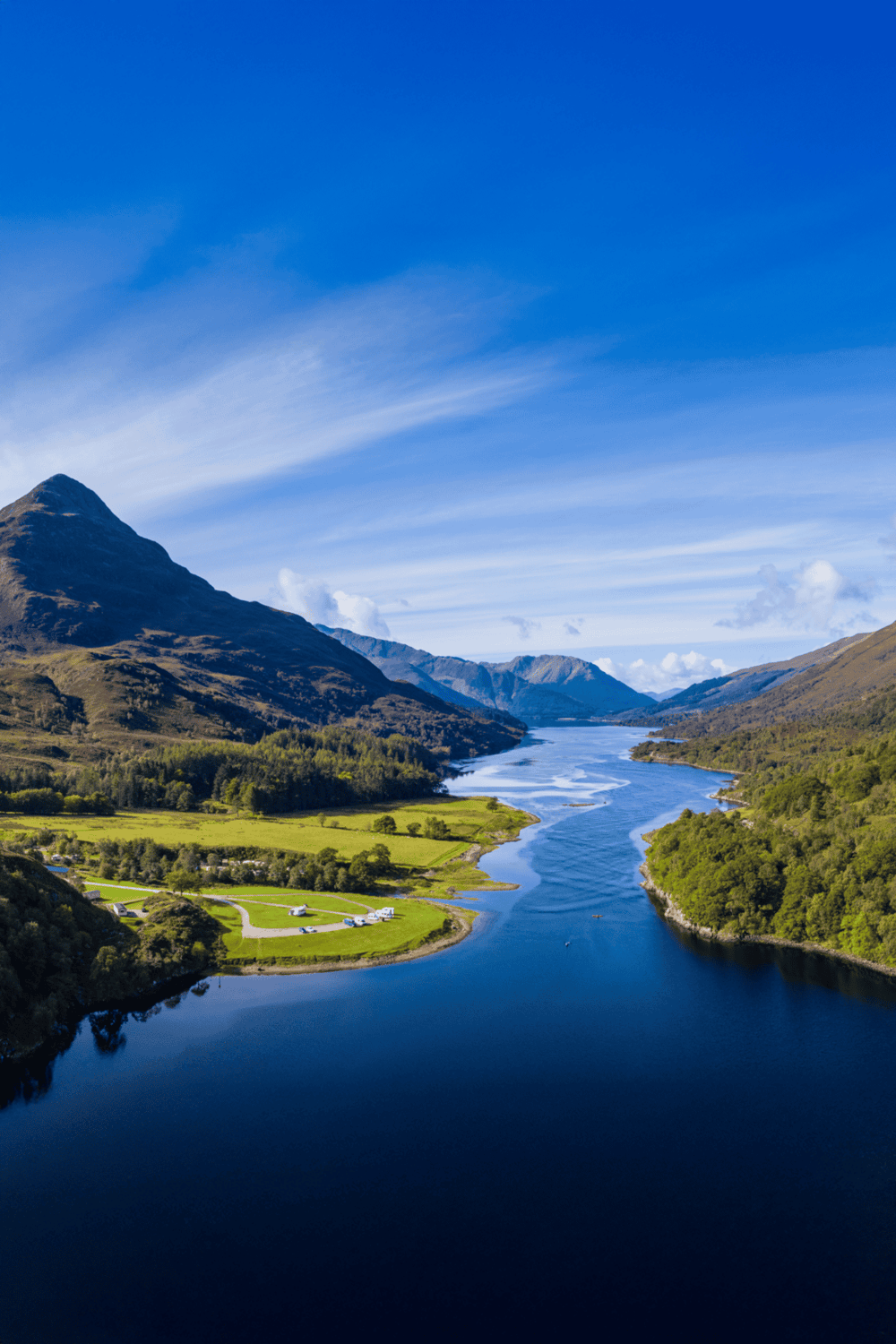 Serene lake and mountain landscape with lush green fields and blue sky, perfect for outdoor adventures.