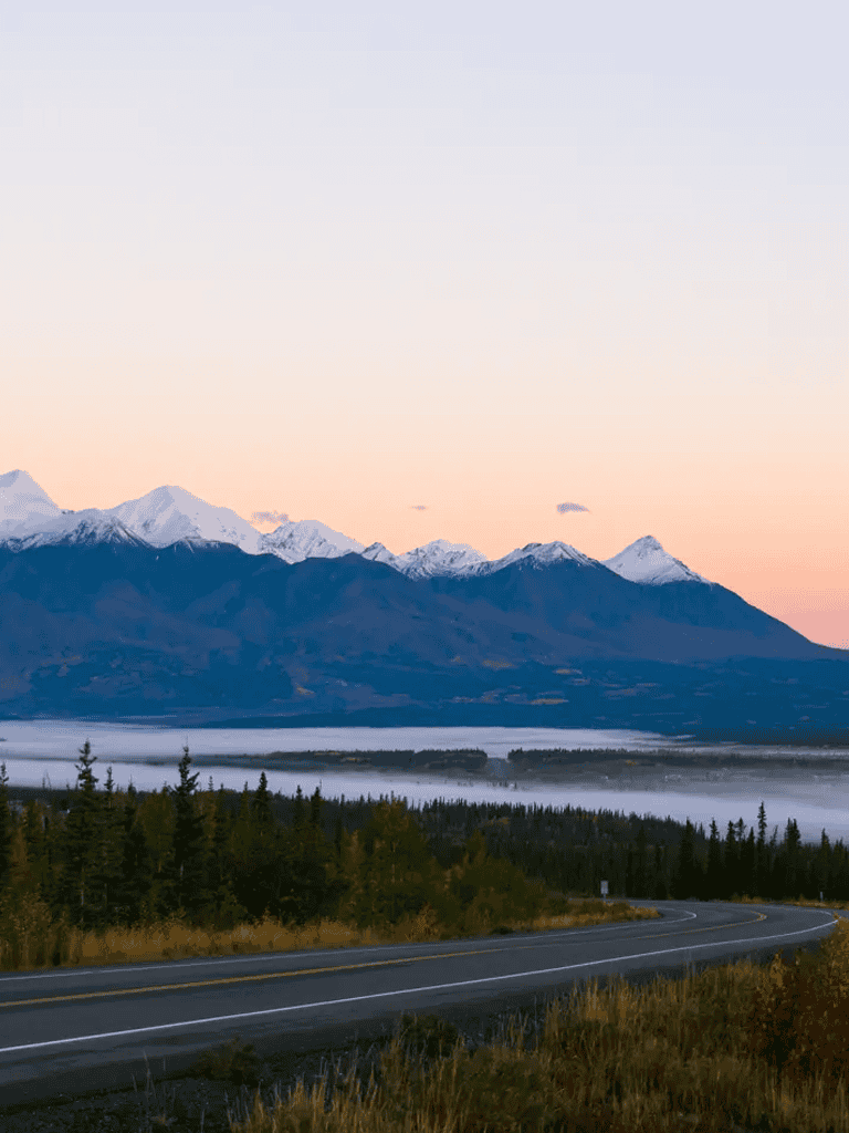 Snow-capped mountains near a winding road in a scenic landscape at sunrise or sunset.