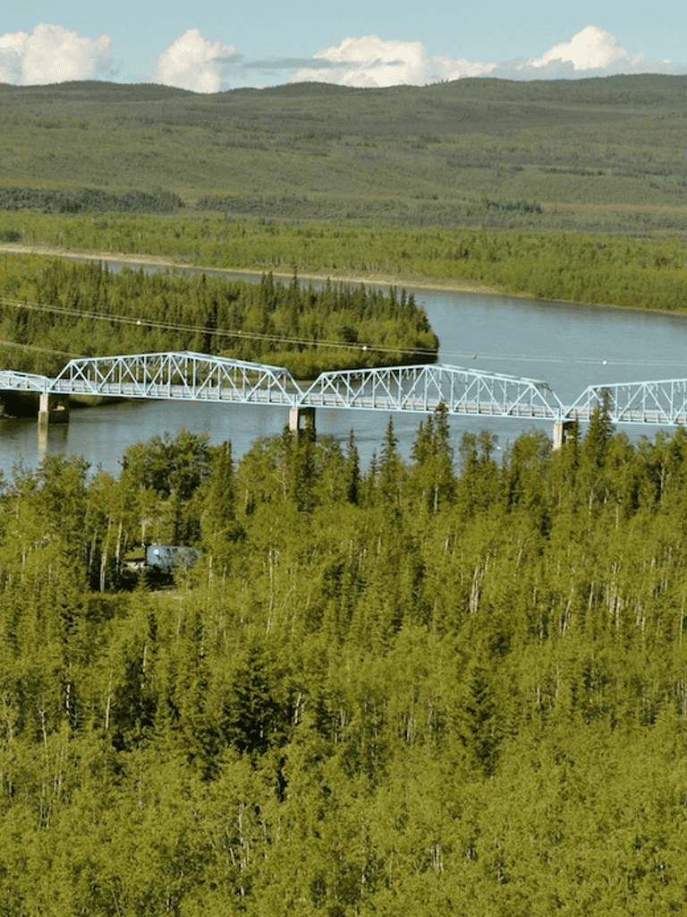 Scenic landscape featuring a bridge over a river in lush green forest with rolling hills in the background.