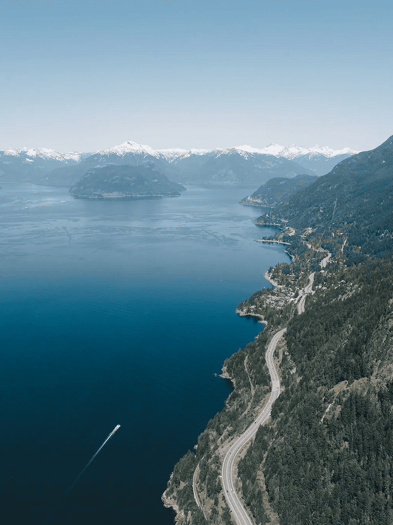 Scenic coastal road along a lake with mountains and snow-capped peaks, stunning natural landscape.