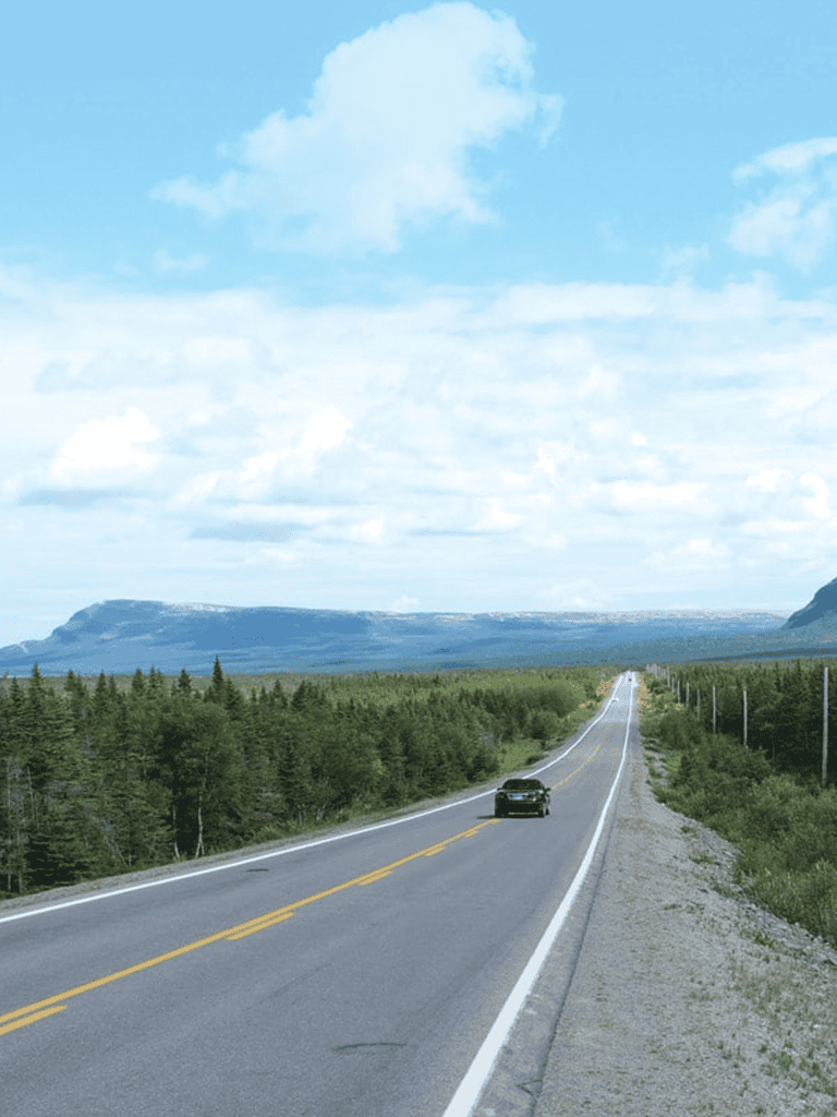 Empty road through lush green forest with mountain views in background, ideal for travel directions.